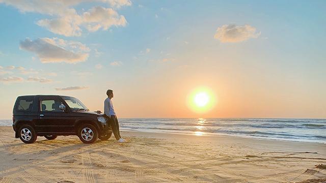 砂浜に止めた車に寄りかかってたたずんでいる人の写真