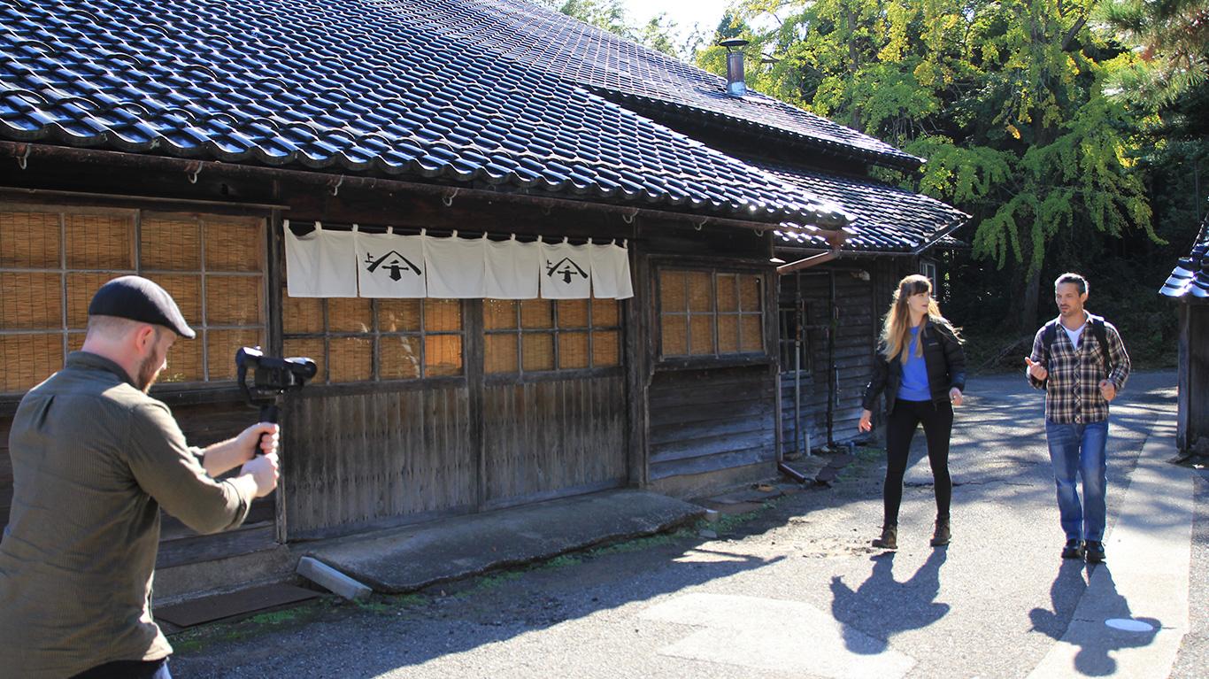 瓦屋根の木造建築の前で歩いている2人とそれを撮影している人の写真