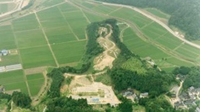 田んぼの中に、木の茂みと古墳群が広がっている写真