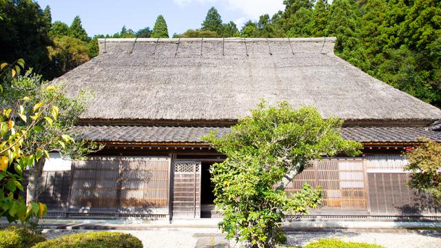 手入れされた庭の木々の後ろにある、扉が少し開いた茅葺屋根の建物の写真