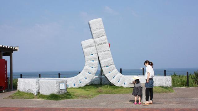 地面からほぼ垂直に反ったモニュメントと、それを見ている親子の写真