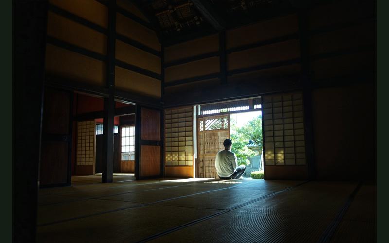 薄暗い和室の中で外を眺めながら胡坐をかいている人の写真