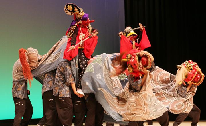 舞台の上で、多くの人が獅子舞や人形の姿を模して演技している写真