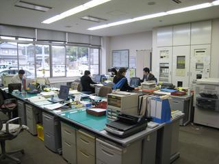 さくらドーム21館内にある事務室で横並びに向かい合いパソコンを使用して仕事に励む4名の職員と一人席で正面を向いて仕事に励む上司の男性を撮影した写真