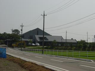 田んぼの奥に、三角形の屋根の大きな施設がある写真
