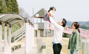 公園で、女の子を高く持ち上げている父親と、横で見ている母親の写真