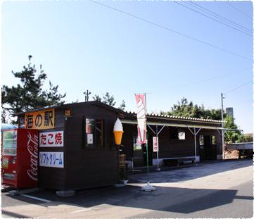 海の駅と書かれた看板が掛けられた小屋の前にのぼりや自動販売機が置かれている写真