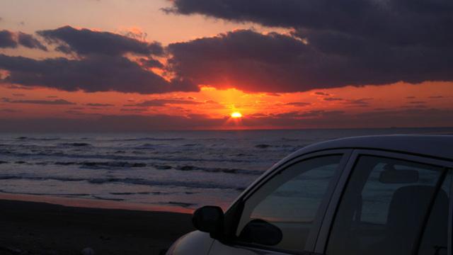 砂浜に止めた車側から日の沈む海の風景を撮影した写真