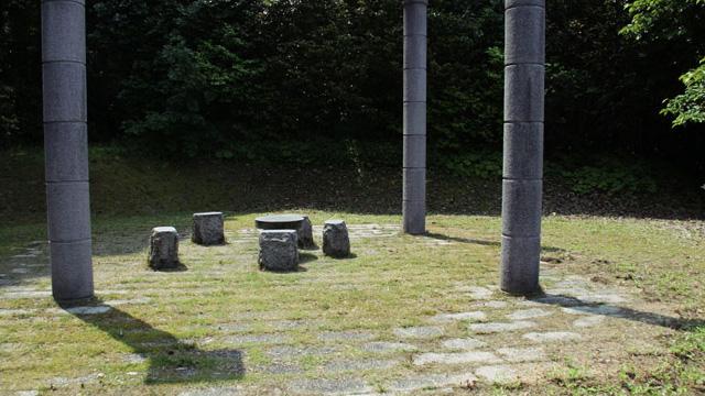 芝生の上に、石のテーブルと椅子を囲むように三本の石柱が立っている写真