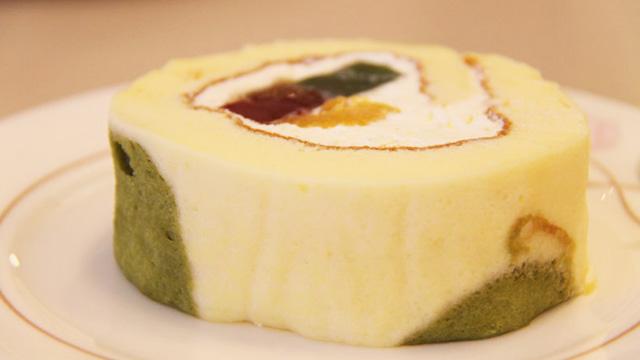 お皿の上に、茶色、緑色、オレンジ色の食材をクリームで包んだロールケーキが置かれている写真