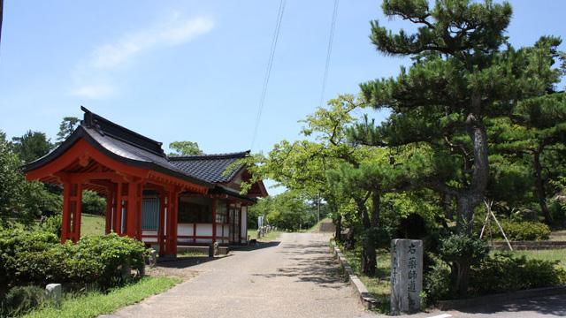 道の左側に朱色の建物があり、右側に松の木などの植栽が生えている写真