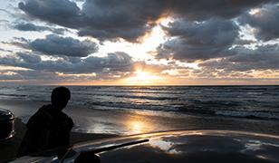 砂浜に止めた車のそばで水平線に重なる太陽を眺める男性の写真(宝達志水町のサイト 千里浜なぎさドライブウェイへのリンク)