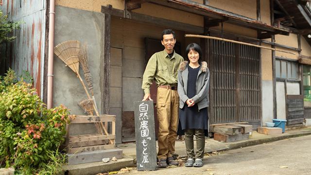 古民家の前で、看板とともに並んでほほ笑んでいる男性と女性の写真