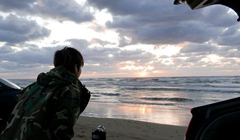 砂浜に止めた車のそばでカメラを海に向けた男性の写真