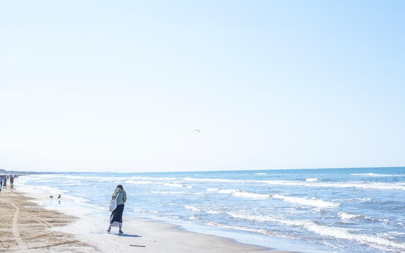 白い鞄を下げた女性が、昼間の浜辺を歩いている写真
