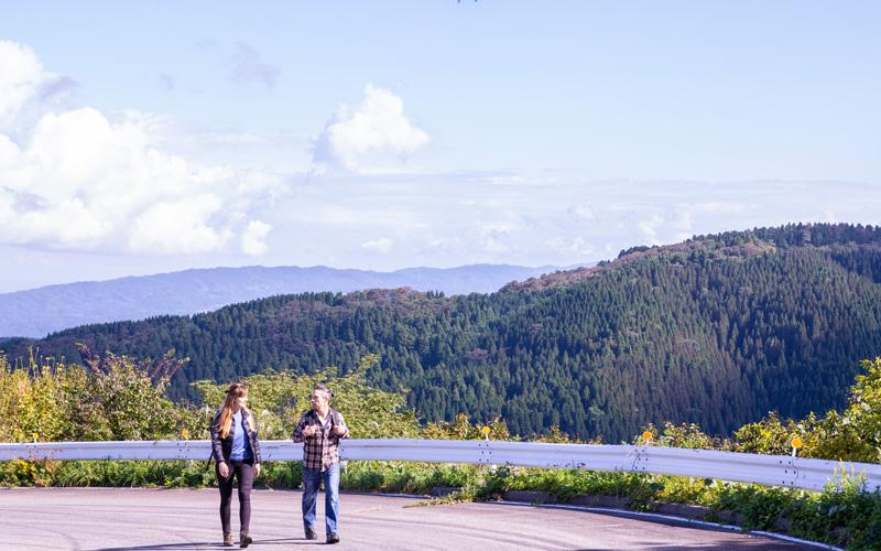 緑の山なみを背に、二人の人が歩いている写真