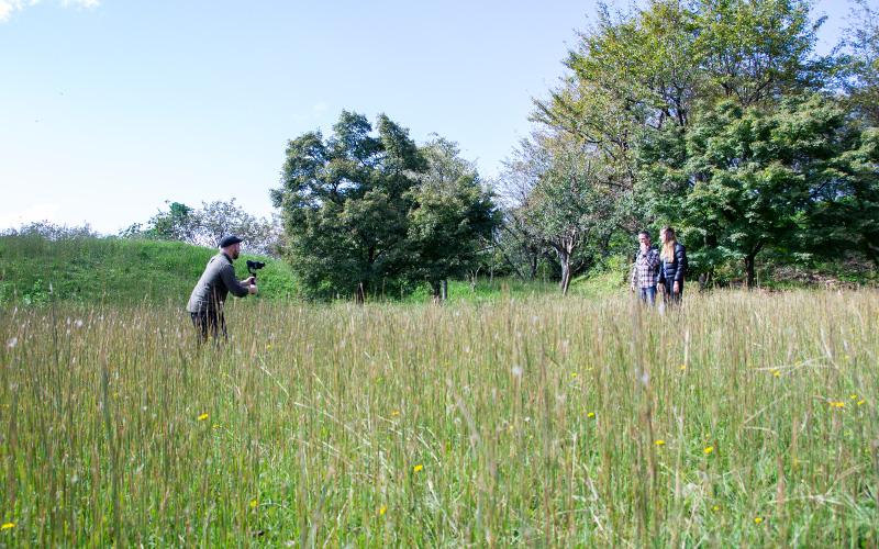 草の生い茂る中で、並んで立っている二人の人にカメラを向けている男性の写真