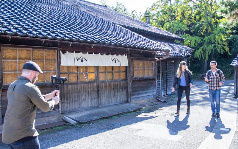 瓦屋根の木造建築の前を歩く2人とその正面からカメラを向ける人の写真