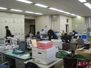 宝達志水町役場内にある税務課事務所でパソコンを使用して仕事に励む男性職員たちの手前で起立して仕事に励む女性職員を正面から撮影した写真