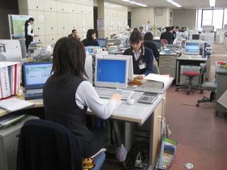 パソコンや書類が並ぶオフィスで仕事をしている職員たちの写真