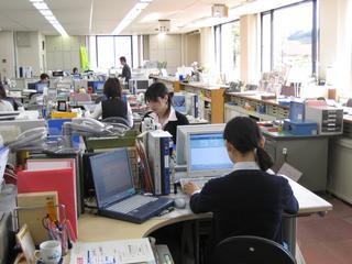 パソコンや書類が置かれたオフィスで仕事をしている職員達の写真