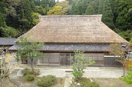 少し色づいた木々と山に囲まれた、茅葺きの住宅の写真