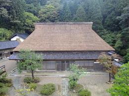 山を背に建つ茅葺きの住宅を、低空から撮影した写真