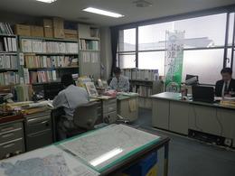 宝達志水町役場内の農林水産課でオフィス用机に向かって黙々と仕事を行う制服姿の職員2人とスーツ姿の上司を撮影した写真